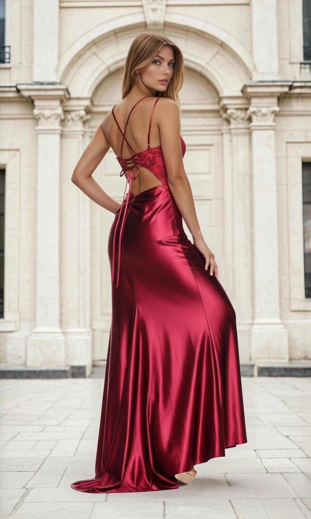 A woman in the Embroidered Corset Long Burgundy Red Prom Dress L633 with thin straps stands outdoors before a light-colored classical building, looking over her shoulder.
